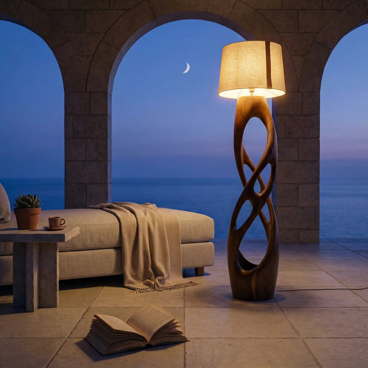 Mediterranean villa terrace at dusk with sculpted wood floor lamp, lounge chair, open book, and sea view.